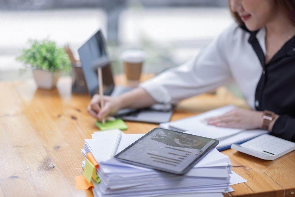 A woman writes on a sticky note while working on paperwork using a laptop on a desk next to a pile of documents with a tablet stacked on top.