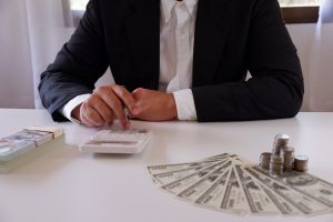 Three business people sit at a table filled with paperwork. One is working on a laptop, another is using a phone, and the third is using a calculator.