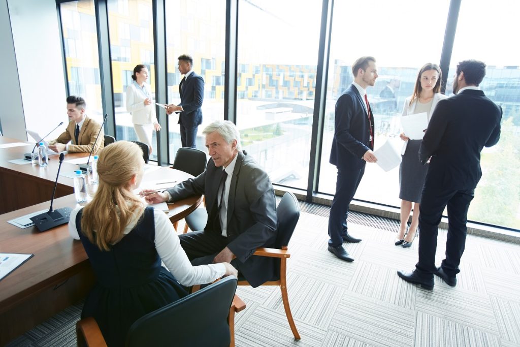 Three businesspeople sit at a large conference table while five others stand talking in groups behind them.