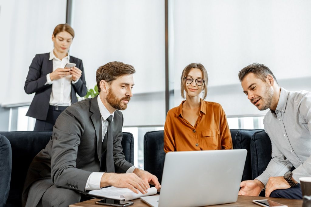 Three businesspeople sit on couches, one showing the others something on a laptop, while an assistant uses a phone in the background.
