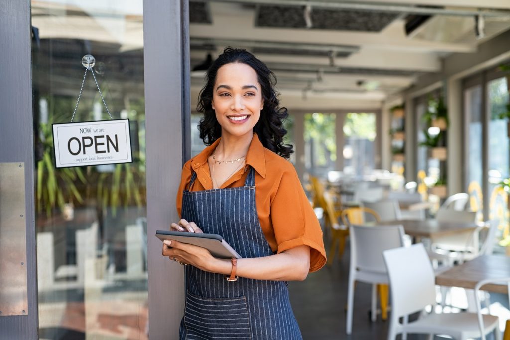 A restaurant owner stands next to the door to her business with an "OPEN" sign hanging from it.